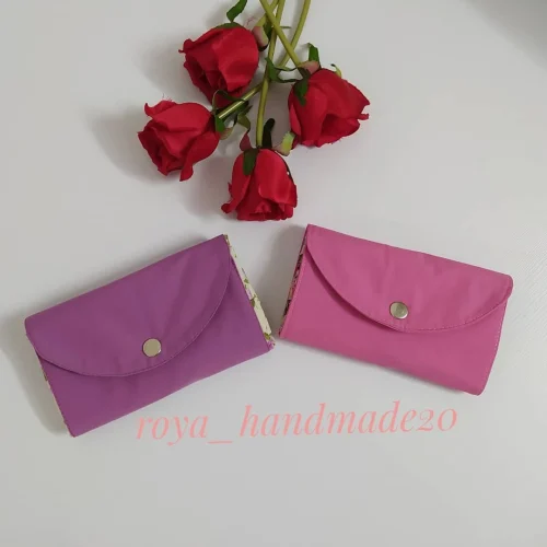 توت بگ تاشو پارچه‌ای در حالت جمع‌شده با دکمه‌ی فشاری، در رنگ‌های بنفش و صورتی، کنار گل رز