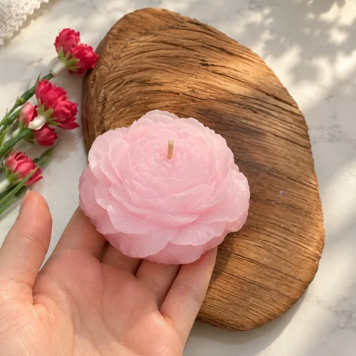شمع دست‌ساز طرح آلاله صورتی در کف دست، کنار تخته‌ی چوبی و گل‌های صورتی