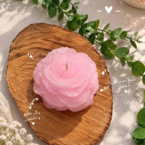 شمع طرح آلاله دست‌ساز به رنگ صورتی روی تخته‌ی چوبی، کنار برگ‌های سبز