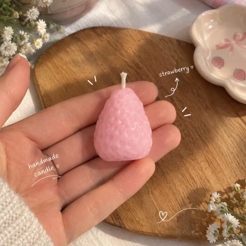 شمع طرح توت‌فرنگی دست‌ساز به رنگ صورتی در کف دست، کنار تخته‌ی چوبی و گل‌های سفید