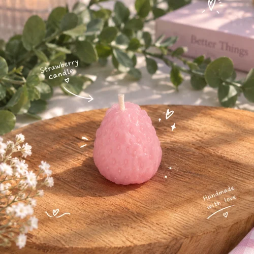 شمع طرح توت‌فرنگی دست‌ساز به رنگ صورتی روی تخته‌ی چوبی، کنار برگ‌های سبز و گل‌های سفید