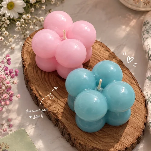 دو شمع طرح روبیک دست‌ساز به رنگ صورتی و آبی روی تخته‌ی چوبی، کنار گل‌های سفید
